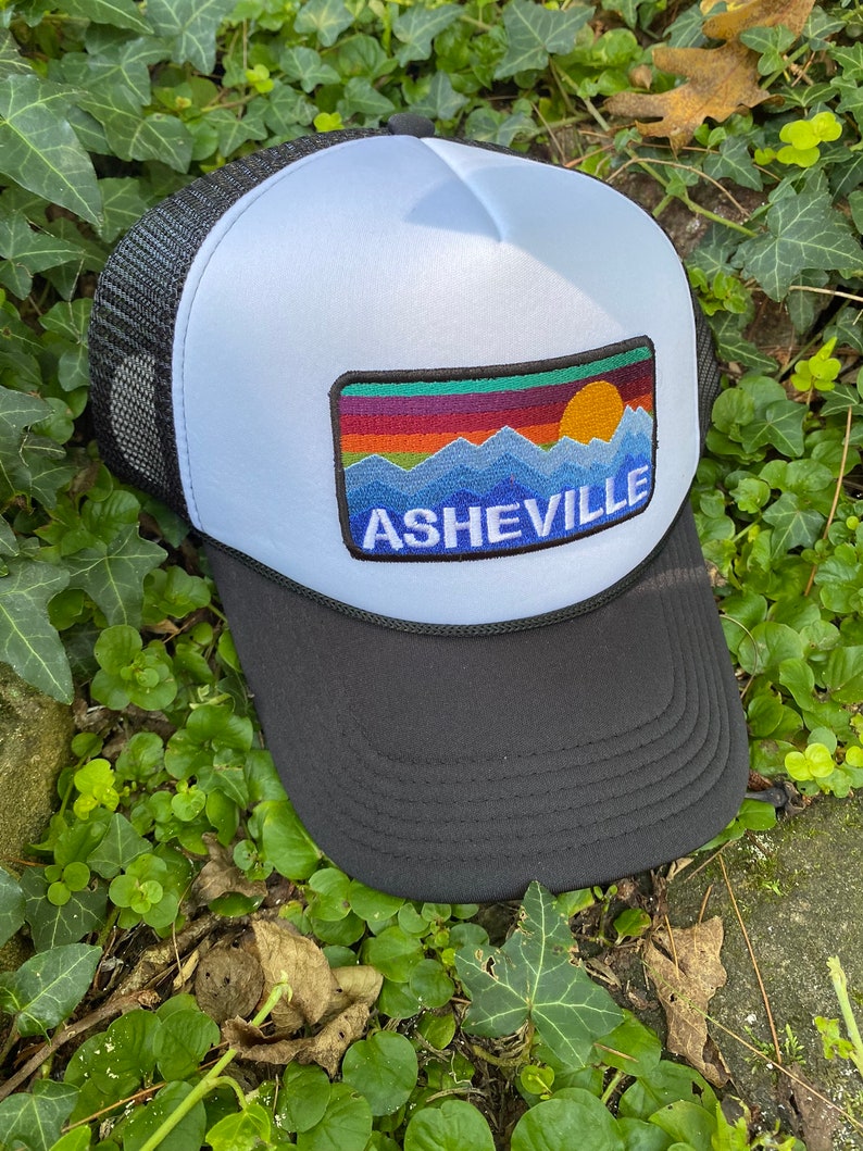Peut inclure: Casquette de camionneur avec une visi&egrave;re et un dos en filet noirs, un panneau avant bleu clair et un &eacute;cusson repr&eacute;sentant une montagne color&eacute;e et un coucher de soleil avec le mot "ASHEVILLE" en lettres blanches. D&eacute;tail de corde noire.