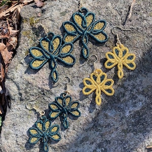Pode incluir: Brincos de trevo bordados em ouro e verde-azulado. Os brincos s&atilde;o exibidos em uma superf&iacute;cie de pedra cinza. Os brincos apresentam um design de trevo de tr&ecirc;s folhas com fio dourado contornando a forma e fio verde-azulado preenchendo.