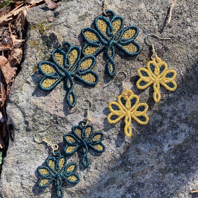 Pode incluir: Brincos de trevo bordados em ouro e verde-azulado. Os brincos s&atilde;o exibidos em uma superf&iacute;cie de pedra cinza. Os brincos apresentam um design de trevo de tr&ecirc;s folhas com fio dourado contornando a forma e fio verde-azulado preenchendo.