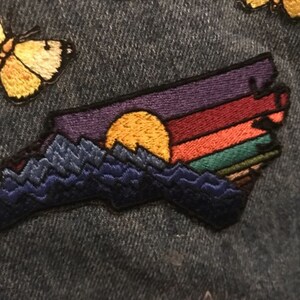 Pode incluir: Patch bordado com um p&ocirc;r do sol sobre uma cordilheira. O design apresenta um sol amarelo e um c&eacute;u em tons de roxo, vermelho, laranja e verde. As montanhas s&atilde;o azul escuro. O patch est&aacute; em um fundo de ganga.