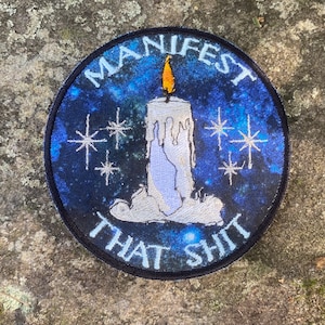 May include: Round patch with a blue galaxy background and a white candle with a yellow flame. The patch has the words "MANIFEST THAT SHIT" in white lettering around the candle and stars.