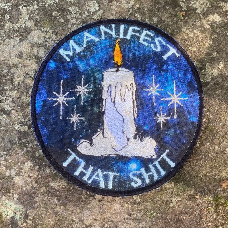 May include: Round patch with a blue galaxy background and a white candle with a yellow flame. The patch has the words "MANIFEST THAT SHIT" in white lettering around the candle and stars.