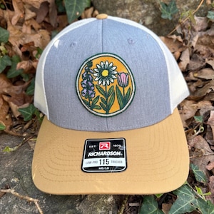 May include: A trucker cap with a grey front panel, white mesh back, and a tan peak. The cap has a circular patch with a floral design in yellow, green, purple, and pink. The Richardson brand label is visible.