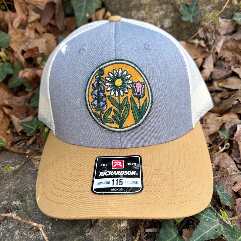 May include: A trucker cap with a grey front panel, white mesh back, and a tan peak. The cap has a circular patch with a floral design in yellow, green, purple, and pink. The Richardson brand label is visible.