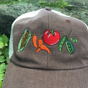May include: A brown baseball cap with a white mesh back. The front of the cap features embroidered images of a corn cob, chili peppers, a tomato, and pea pods. The cap has a curved brim and a button on top.