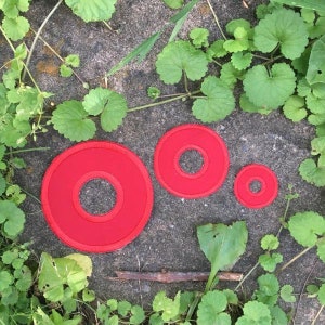May include: Three red circular patches with a hole in the center are arranged on a gray stone surface, surrounded by green plants. The patches vary in size, with the largest on the left and the smallest on the right.