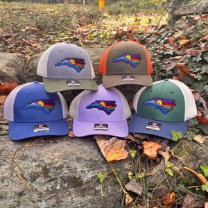 K&ouml;nnte beinhalten: Sechs Trucker-Caps in verschiedenen Farben, darunter Blau, Gr&uuml;n, Lila und Olivgr&uuml;n. Jede Kappe zeigt eine Umrisslinie des Bundesstaates North Carolina mit einem Berg- und Sonnenuntergangsdesign. Die Kappen haben Mesh-R&uuml;cken und verstellbare Verschl&uuml;sse.