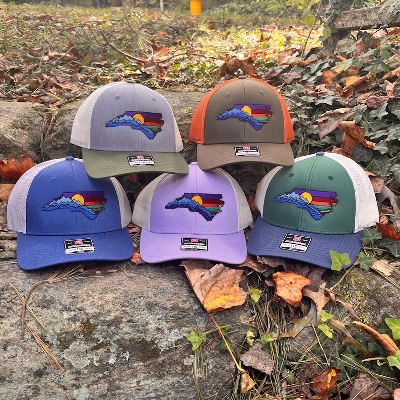 K&ouml;nnte beinhalten: Sechs Trucker-Caps in verschiedenen Farben, darunter Blau, Gr&uuml;n, Lila und Olivgr&uuml;n. Jede Kappe zeigt eine Umrisslinie des Bundesstaates North Carolina mit einem Berg- und Sonnenuntergangsdesign. Die Kappen haben Mesh-R&uuml;cken und verstellbare Verschl&uuml;sse.
