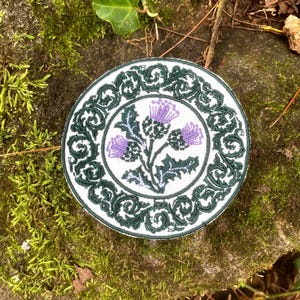 May include: Round embroidered patch featuring a purple thistle design on a white background. The thistle is surrounded by green leaves and a green decorative border. The patch is likely for clothing or accessories.
