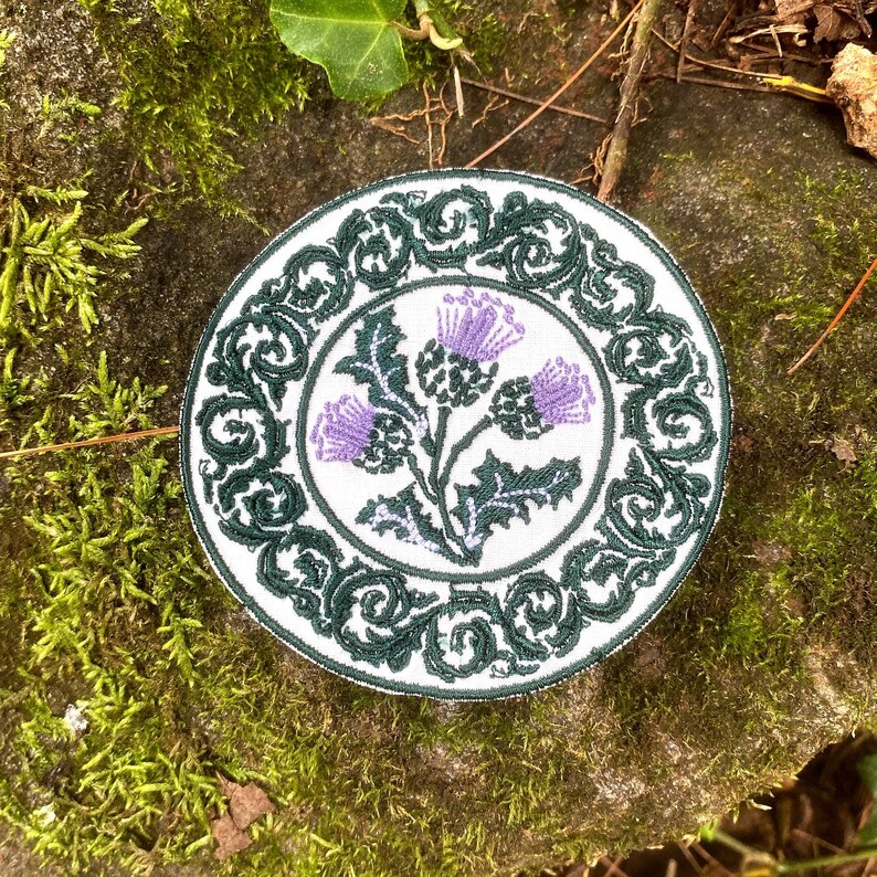 May include: Round embroidered patch featuring a purple thistle design on a white background. The thistle is surrounded by green leaves and a green decorative border. The patch is likely for clothing or accessories.