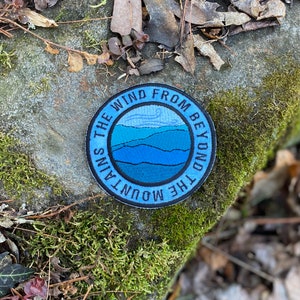 May include: A circular blue patch with the text "THE WIND FROM BEYOND THE MOUNTAINS" around a central design. The design shows layered blue mountain shapes and a wind graphic. The patch is on a moss-covered, natural background.