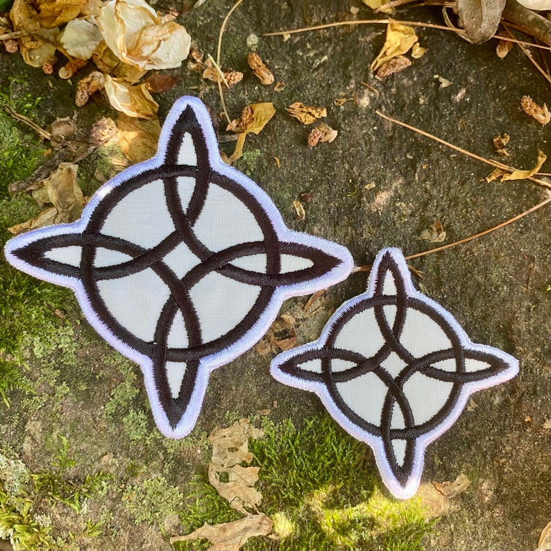 May include: Two white and black Celtic knot patches. The patches feature a black knot design with a white background and a white border. The patches are in different sizes and are placed on a mossy, textured surface.