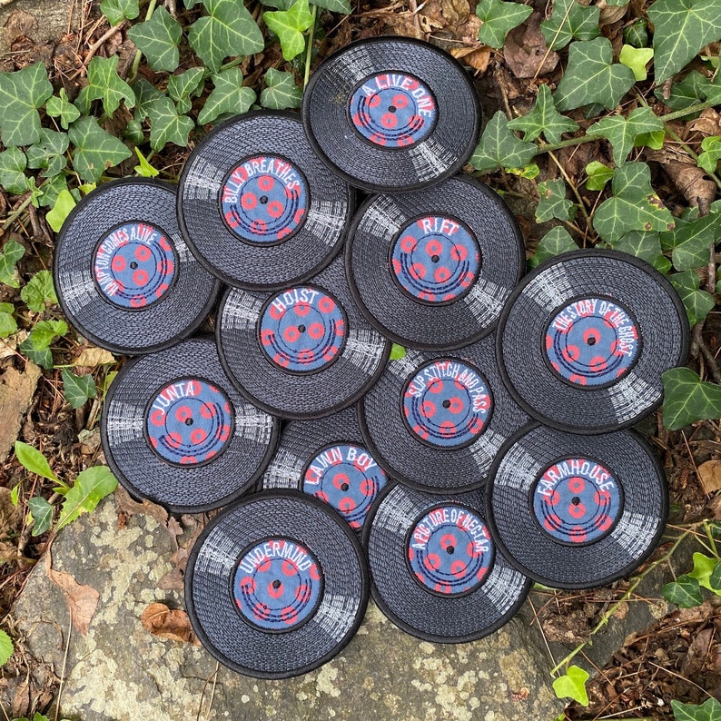 May include: A collection of black, circular coasters designed to resemble vinyl records. Each coaster features a unique title in white text on a blue and red center. The coasters are arranged on a bed of green leaves and a stone.