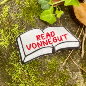 May include: A white patch in the shape of an open book with the words "READ VONNEGUT" embroidered in red. The patch has a black border and is set against a mossy background with green leaves.
