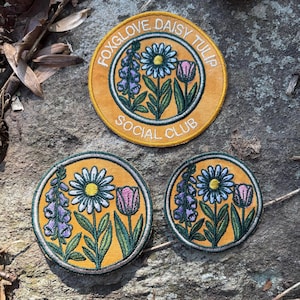 May include: Three embroidered patches with floral designs. The largest patch has the text "FOXGLOVE DAISY TULIP SOCIAL CLUB" around the edge. The patches feature daisies, tulips, and foxgloves in shades of yellow, green, purple, and pink.