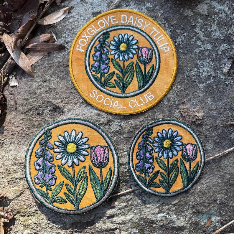 Pu&ograve; includere: Tre toppe ricamate con disegni floreali. La toppa pi&ugrave; grande ha il testo "FOXGLOVE DAISY TULIP SOCIAL CLUB". Le toppe presentano margherite, tulipani e digitali nei toni del giallo, verde, viola e rosa.