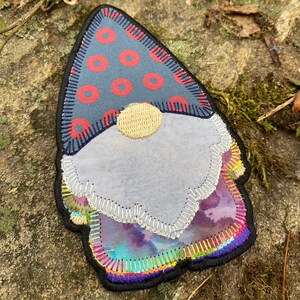 May include: A fabric gnome patch with a blue hat featuring red polka dots. The gnome has a light blue face and a beige nose. The gnome's beard and clothing are a mix of colors, including purple, yellow, and green, with a black border.