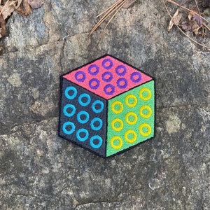 May include: A colorful cube-shaped patch with a black border. Each face of the cube is a different color: pink, green, black, and the other faces are not visible. Each face has a pattern of circular designs in contrasting colors.