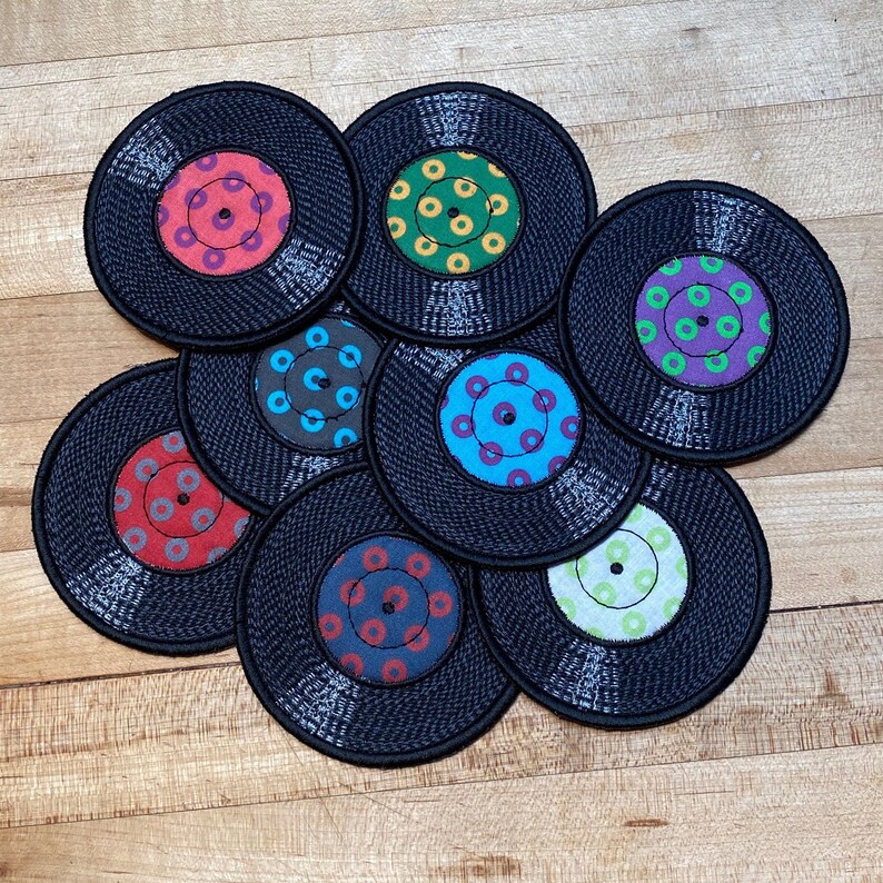 Pu&ograve; includere: Una collezione di nove sottobicchieri neri a forma di vinile con etichette centrali colorate. Le etichette presentano vari motivi circolari nei toni del rosso, blu, verde, viola e grigio. I sottobicchieri sono disposti su una superficie di legno.