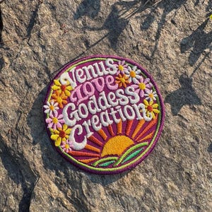 May include: Circular embroidered patch with the words "Venus Love Goddess Creation". The patch has a purple border, a yellow and orange sun, and colorful flowers. The lettering is white and pink.