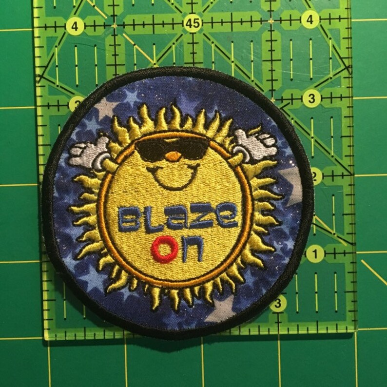 Puede incluir: Parche bordado con un sol sonriente con gafas de sol. El sol es amarillo con un contorno dorado y llamas radiantes. El parche tiene las palabras "Blaze On" en azul y rojo. El fondo es azul con estrellas blancas.