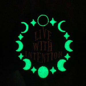 May include: A dark background with a glowing green design. The design includes crescent moons, stars, and a circle, encircling the reddish-brown text "LIVE WITH INTENTION".