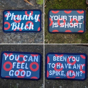 May include: Four rectangular embroidered patches with dark blue borders. Each patch has a teal background with red circle patterns. The patches feature white text: "Phunky Bitch", "Your Trip Is Short", "You Can Feel Good", and "Been You To Have Any Spike, Man?"