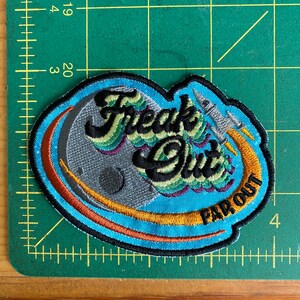 May include: Embroidered patch with the words "Freak Out" and "Far Out" in black, green, and purple. The patch features a gray moon, a rocket ship, and a blue and orange design. The patch is approximately 3 inches wide.