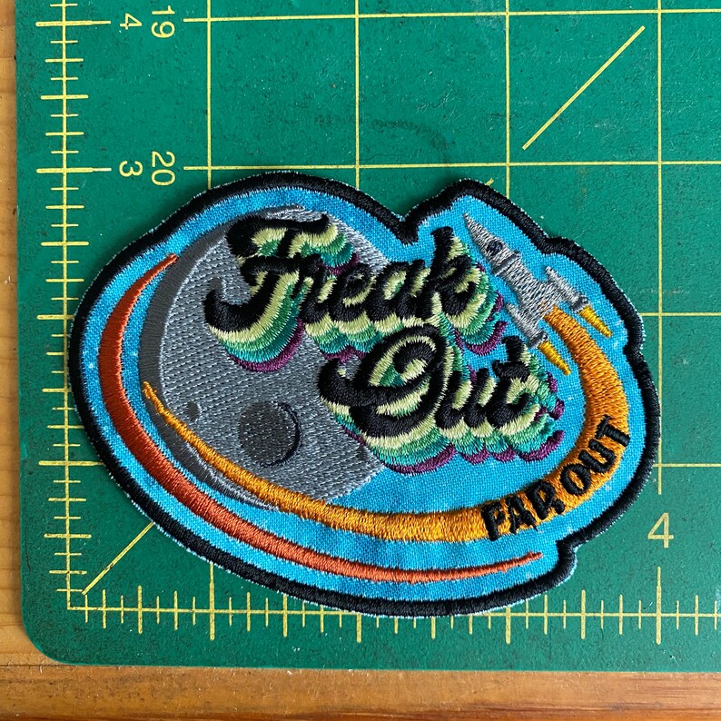 May include: Embroidered patch with the words "Freak Out" and "Far Out" in black, green, and purple. The patch features a gray moon, a rocket ship, and a blue and orange design. The patch is approximately 3 inches wide.