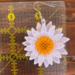 May include: A single daisy-shaped earring with a white lace petal design and a yellow center. The earring has a silver-colored hook. The earring is approximately 2 inches in diameter.