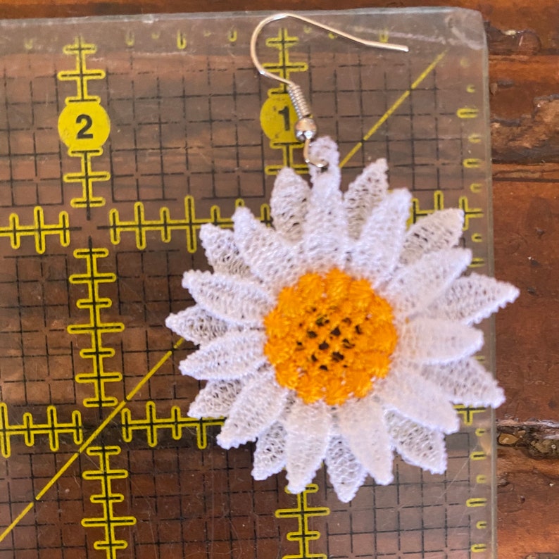 May include: A single daisy-shaped earring with a white lace petal design and a yellow center. The earring has a silver-colored hook. The earring is approximately 2 inches in diameter.