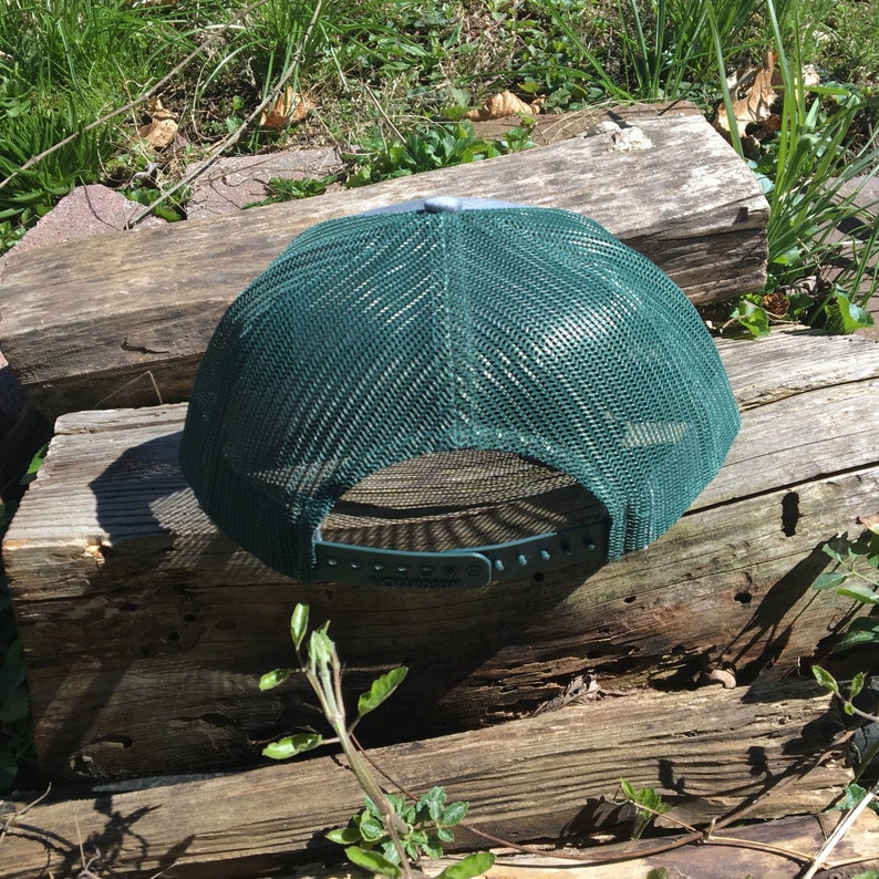 May include: A green trucker hat with a mesh back and a gray top. The hat is sitting on a weathered wooden surface, with green grass and foliage in the background. The hat has an adjustable snap closure.