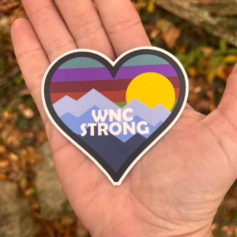 Puede incluir: Pegatina en forma de coraz&oacute;n con un dise&ntilde;o de monta&ntilde;a y puesta de sol. La pegatina presenta una paleta de colores azul, morado, rojo y amarillo. El texto "WNC STRONG" est&aacute; impreso en blanco sobre un fondo azul.