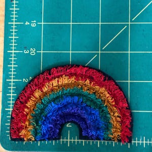 May include: A colorful rainbow patch with red, orange, yellow, green, and blue threads. The patch is approximately 3 inches wide and 1.5 inches tall. The rainbow is embroidered on a black background.