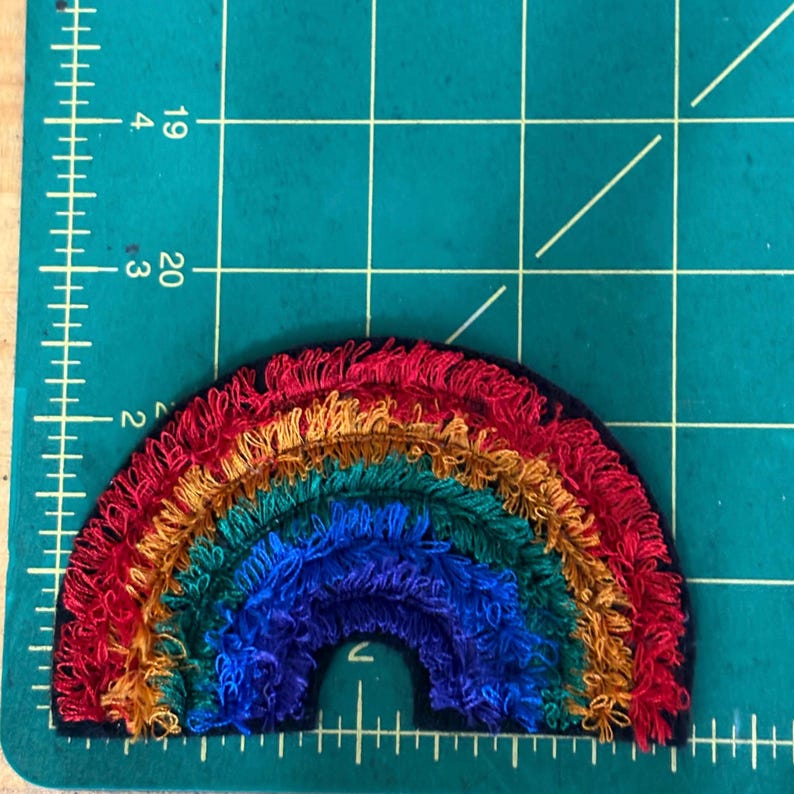 May include: A colorful rainbow patch with red, orange, yellow, green, and blue threads. The patch is approximately 3 inches wide and 1.5 inches tall. The rainbow is embroidered on a black background.