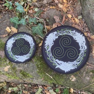 May include: Two black circular patches with embroidered designs. Each patch features a white wolf head on either side of a green triple spiral symbol, surrounded by a green vine-like border. The patches are set on a stone surface.