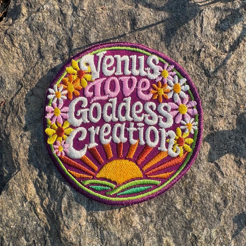May include: Embroidered patch with the words "Venus Love Goddess Creation" in white script. The patch has a purple border and features a sunrise design with yellow and orange rays. Floral accents in yellow and pink surround the text.