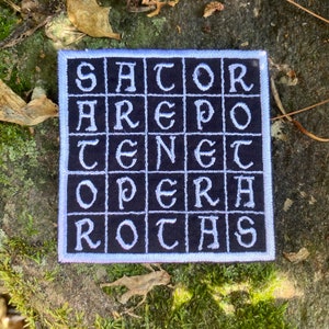 May include: Square patch with the text "SATOR AREPO TENET OPERA ROTAS" in white, block letters on a black background. The patch has a white border and is set against a natural, mossy background.