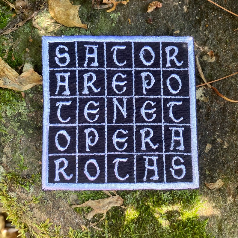 May include: Square patch with the text "SATOR AREPO TENET OPERA ROTAS" in white, block letters on a black background. The patch has a white border and is set against a natural, mossy background.