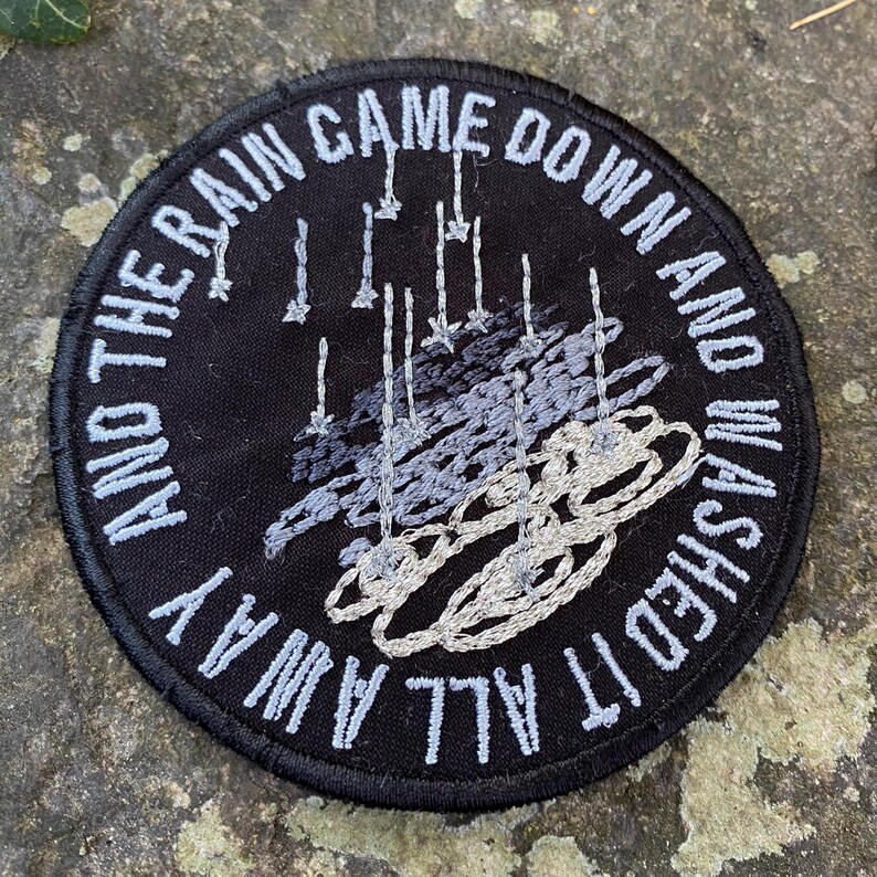 Pode incluir: Patch redondo bordado em preto com o texto branco "AND THE RAIN CAME DOWN AND WASHED IT ALL AWAY". O patch apresenta um design bordado cinza e branco de chuva caindo.