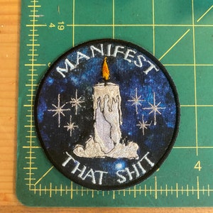 May include: Round embroidered patch with a dark blue starry background and a white candle with a yellow flame. The text "MANIFEST THAT SHIT" is arched around the top and bottom of the patch in white lettering.