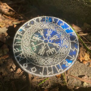 May include: Circular patch with a silver embroidered border and a blue and green centre. The design features a central symbol surrounded by runes. The patch is round and appears to be made of fabric.