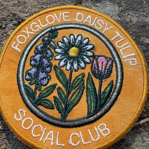 Pu&ograve; includere: Toppa rotonda ricamata con un disegno floreale. La toppa ha uno sfondo arancione e un testo bianco che recita "FOXGLOVE DAISY TULIP SOCIAL CLUB". Il disegno presenta una margherita, un tulipano e una digitale in tonalit&agrave; di viola, bianco, rosa e verde.