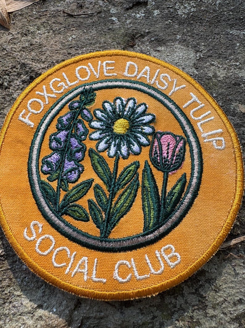 Pu&ograve; includere: Toppa rotonda ricamata con un disegno floreale. La toppa ha uno sfondo arancione e un testo bianco che recita "FOXGLOVE DAISY TULIP SOCIAL CLUB". Il disegno presenta una margherita, un tulipano e una digitale in tonalit&agrave; di viola, bianco, rosa e verde.