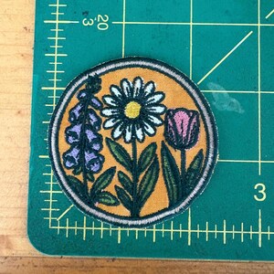 Pu&ograve; includere: Toppa ricamata circolare con un design floreale. La toppa ha uno sfondo giallo e un bordo argentato. Il design include una margherita bianca, un tulipano rosa e fiori di digitale viola con foglie verdi.
