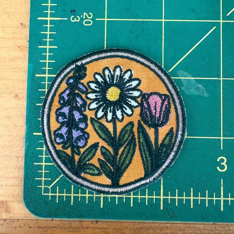 Pu&ograve; includere: Toppa ricamata circolare con un design floreale. La toppa ha uno sfondo giallo e un bordo argentato. Il design include una margherita bianca, un tulipano rosa e fiori di digitale viola con foglie verdi.