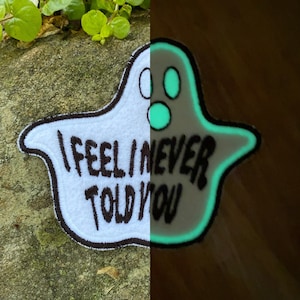 May include: A white ghost-shaped patch with the text "I FEEL I NEVER TOLD YOU" in black. The patch has a brown border and glows green in the dark. The patch is on a stone surface.