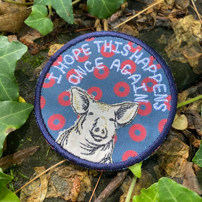 May include: Round patch with a blue background featuring a pig illustration and the text "I HOPE THIS HAPPENS ONCE AGAIN" in white. The pig is white with black details, and the background has red circles.