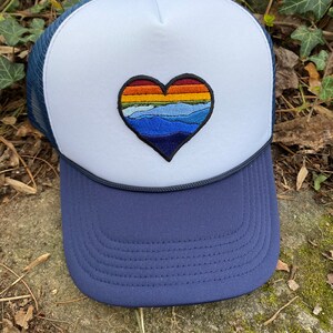 May include: A navy and white trucker hat with a heart-shaped embroidered design. The heart features a colourful landscape of mountains and a sunset. The hat has a mesh back and a curved brim, suitable for outdoor activities.