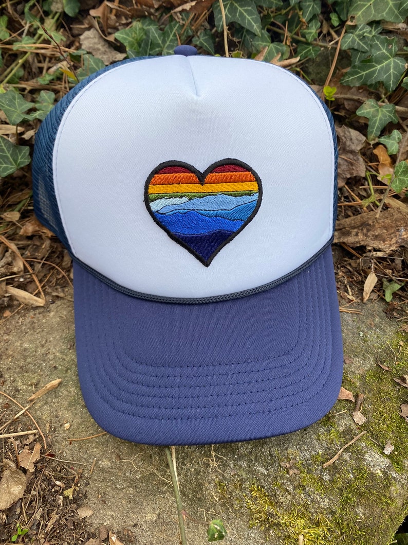 May include: A navy and white trucker hat with a heart-shaped embroidered design. The heart features a colourful landscape of mountains and a sunset. The hat has a mesh back and a curved brim, suitable for outdoor activities.